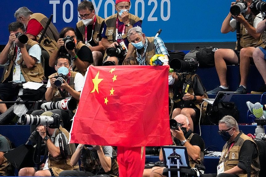 A person unfurls a large Chinese flag at a stadium, many photographers are seen in the background