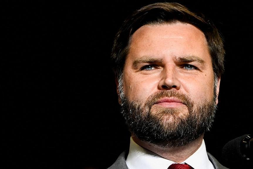 A man with full beard and moustache stands agaainst a black background