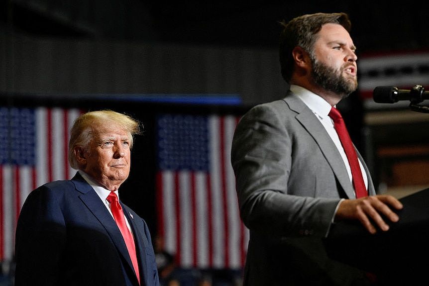 JD Vance speaks at a lectern as Donald Trump stands behind watching, with two US flags