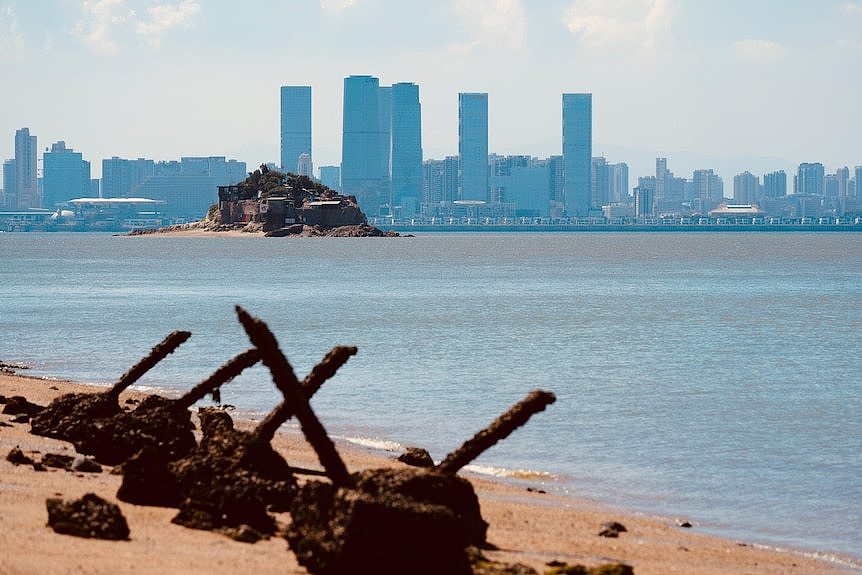 A small island sits in the centre of the sea with the outline of a city behind it.