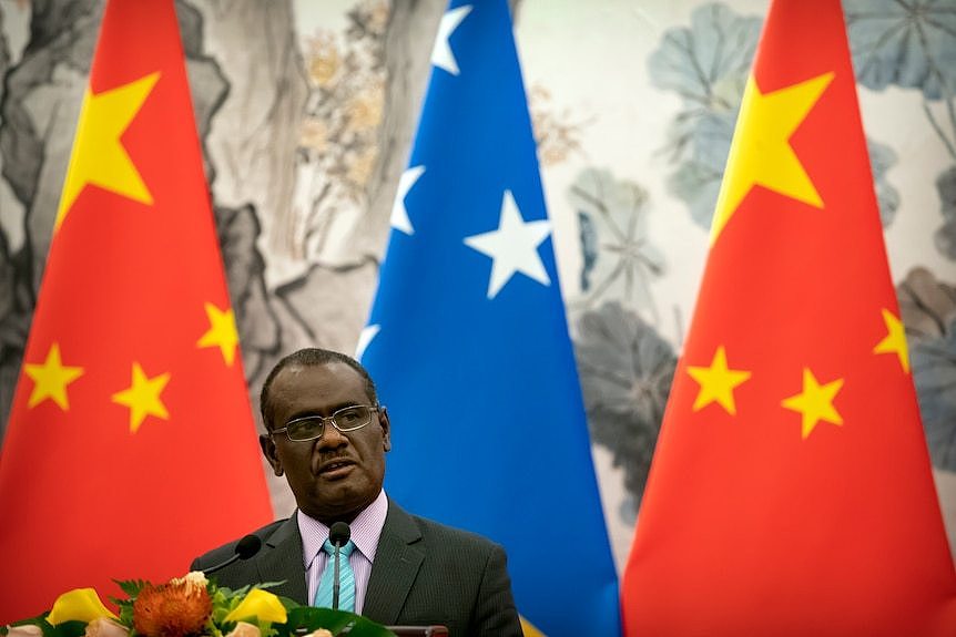 A man in a suit and glasses stands in front of Chinese and Solomon Islands flags.