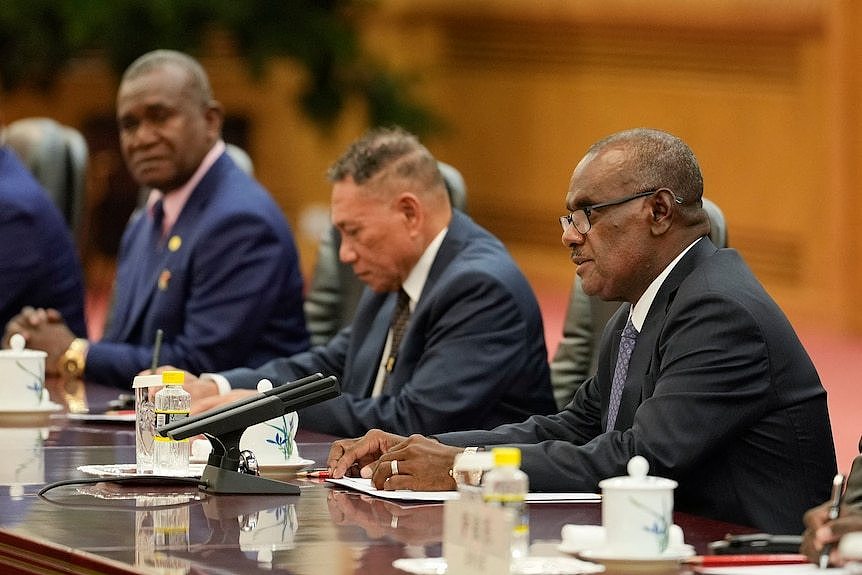 Three men in suits at a table. The man in the foreground is speaking into a microphone.