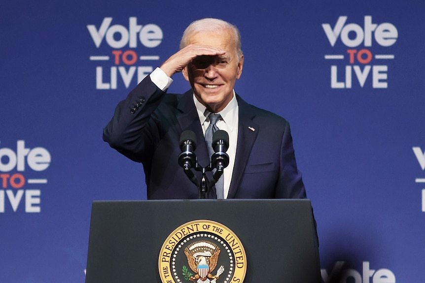 Joe Biden shades his eyes with his right hand as he gives a speech from behind a presidential lectern.