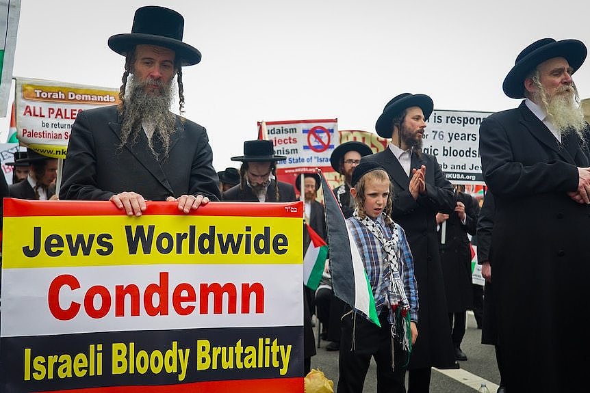 A group of men in Orthodox Jewish attire with a sign saying 'Jews worldwide condemn Israeli bloody brutality'.