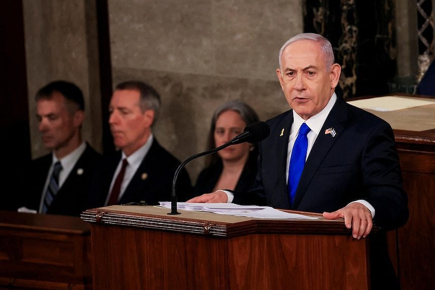 Benjamin Netanyahu wearing a blue tie stands at a lectern speaking into a microphone