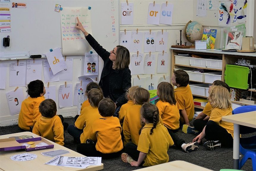 twelve students sit on the mat looking at teacher who is pointing at whiteboard.