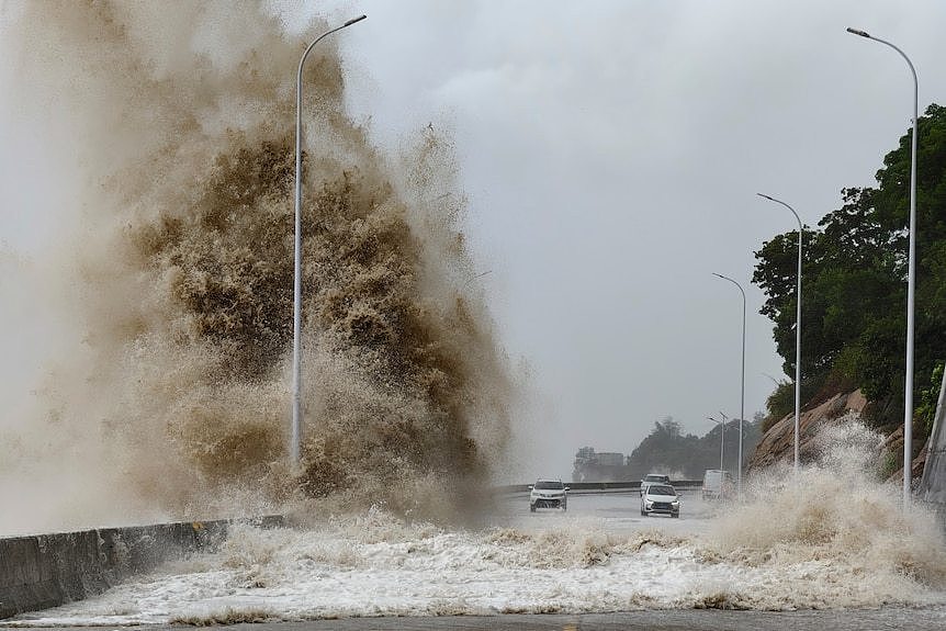 Waves rise high after striking a seawall next to a road, as cars pass