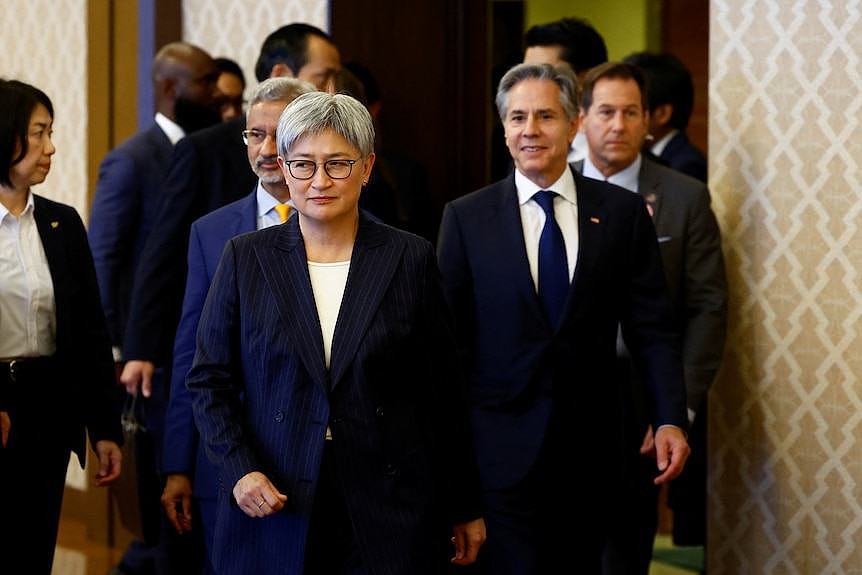 A woman in a suit with short white hair walks forward, flanked by two men in suits.