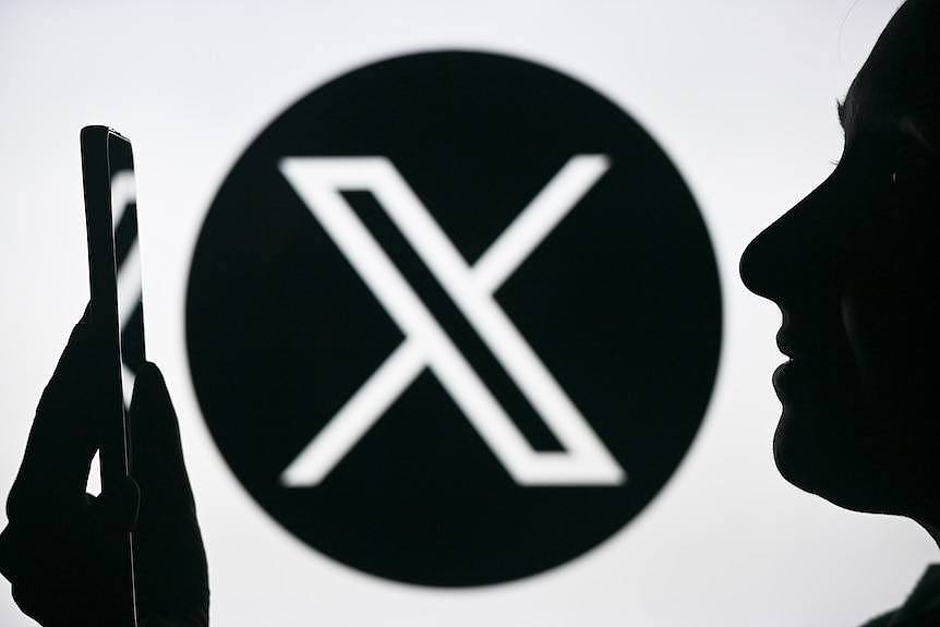 The logo of tech company X on a wall, with the profile of a person looking at a smartphone in front of it.