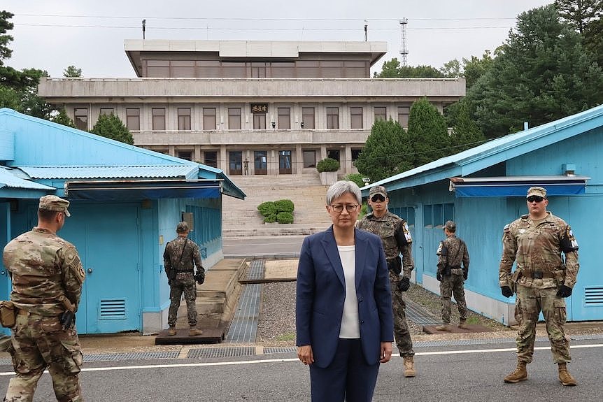 Penny Wong stands in a blue suit looking stern surrounded by soldiers in camouflage in front of a large building