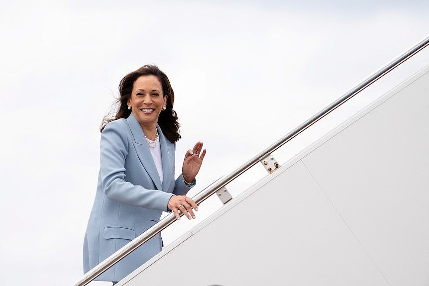 Kamala Harris, wearing a light blue suit, walks up an aircraft staircase and waves.