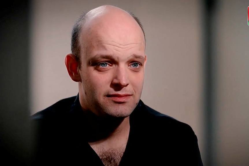 A partially bald man wearing a black shirt stares to the side of the camera.