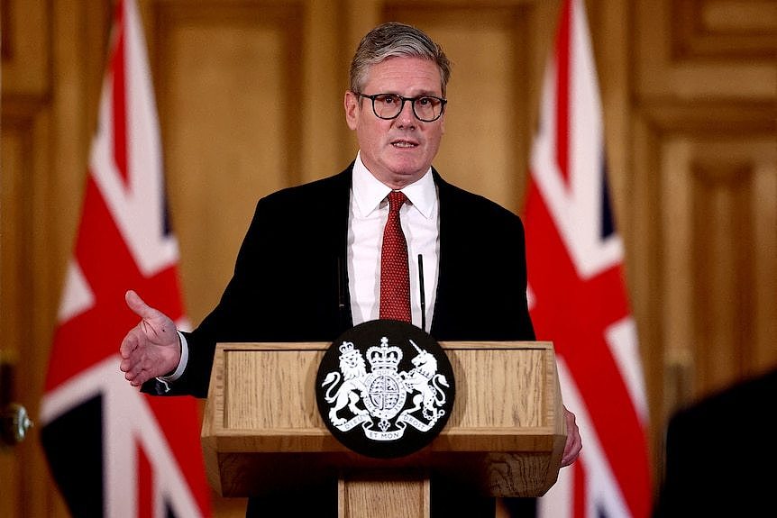 Keir Starmer stands at a podium, with two British flags in the background.