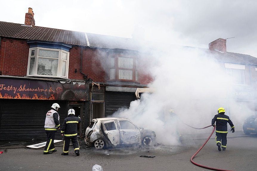 Three firefighters surround a burnt-out car, a plume of smoke rising from the scene.