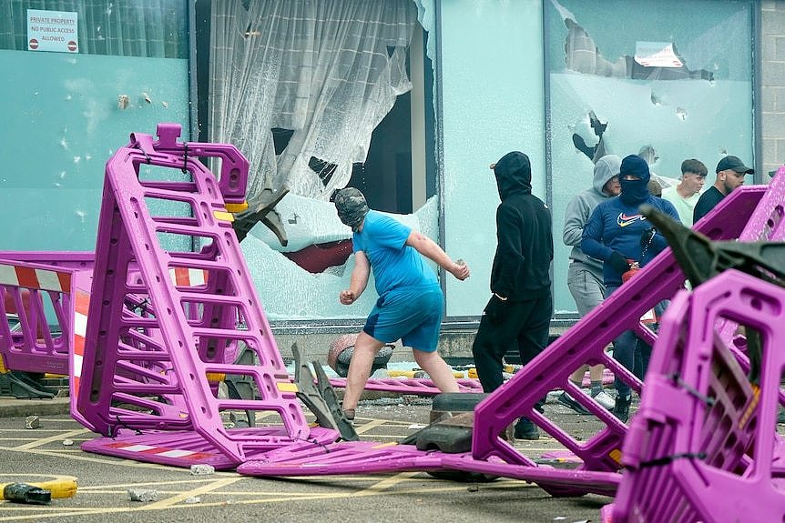 A man throws an object through a window, behind a set of pink barriers.