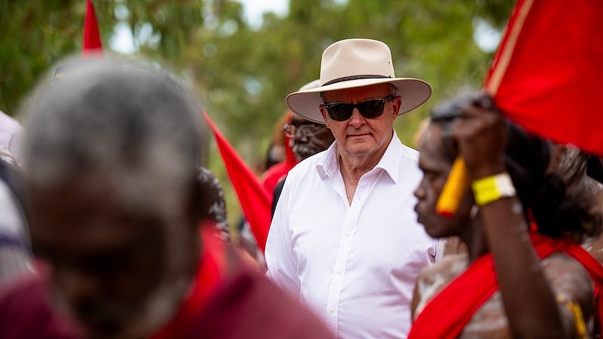 安东尼·阿尔巴尼斯 (Anthony Albanese) 戴着帽子和太阳镜走在一群穿着红色服装、涂着人体彩绘的土著男子中间。