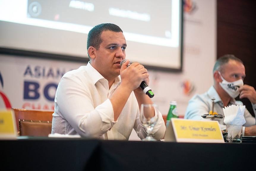 A man sits holding a microphone at a table and is talking