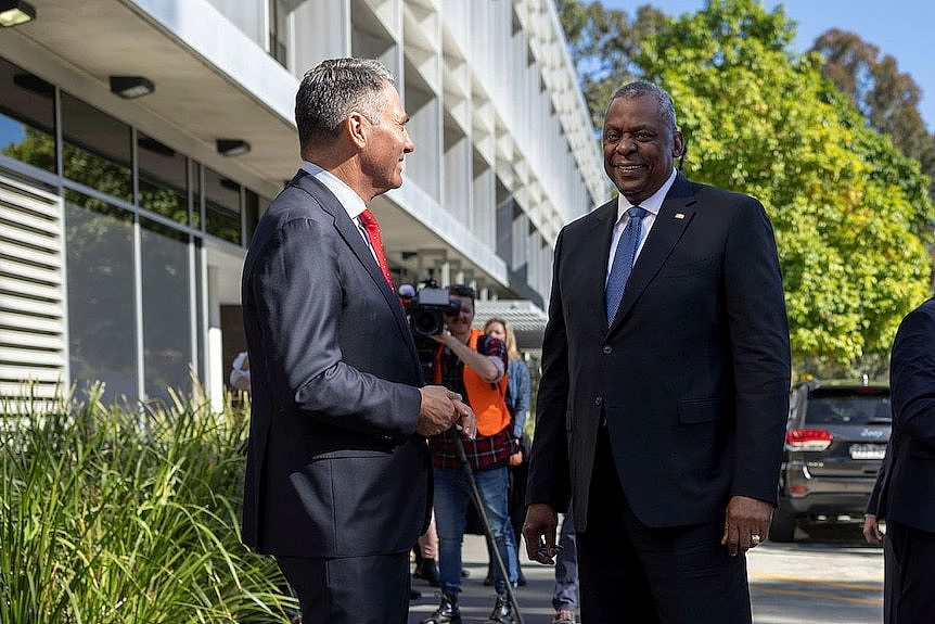 Two middle-aged men in suits stand outside a building in sunshine with camera operators behind