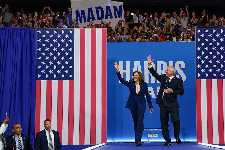 Kamala Harris and Tim Walz wave to a large crowd from a stage, with the words 'Harris Walz' on a wall behind them.