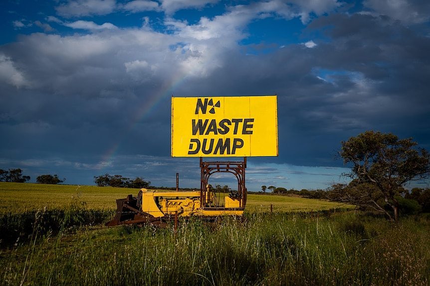 A tractor with a sign that reads 'No Waste Dump' is parked in a wheat field. A rainbow is in the sky.