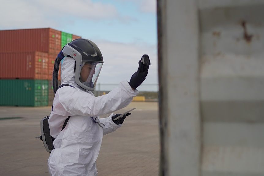 A worker in protective gear testing radiation levels around a shipping container.