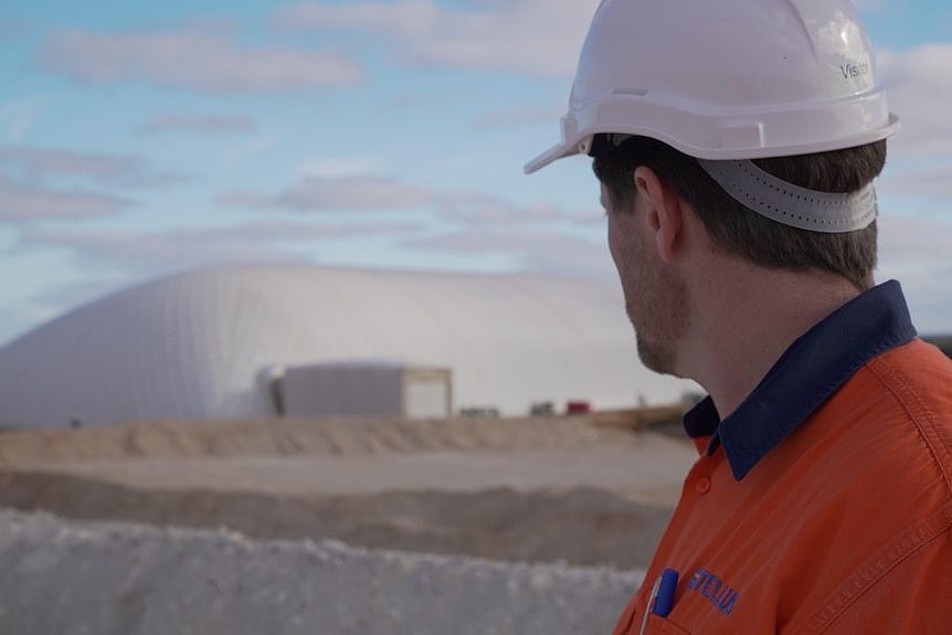 A man wearing a helmet and high-vis looks back at a large white building. 