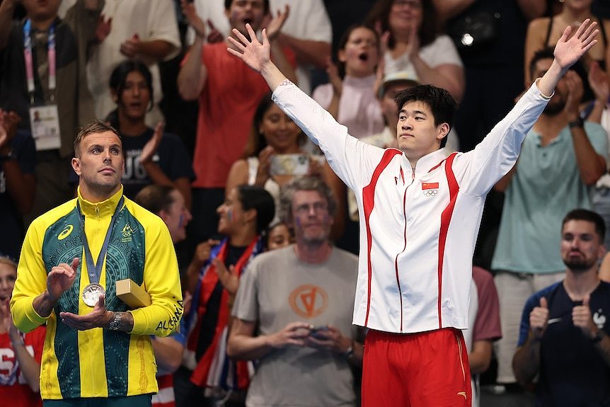 A man celebrates winning a gold medal with his arms wide on the top of the podium 