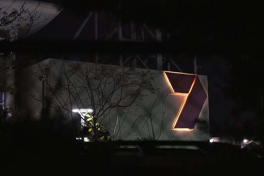 A Channel 7 building can be seen through a fence at night. It has an illuminated 7 logo on its wall.