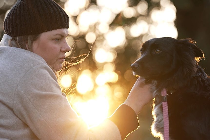 A woman with a neutral expression touches the face of a dog. Between them the setting sun is shining through leaves of a tree.