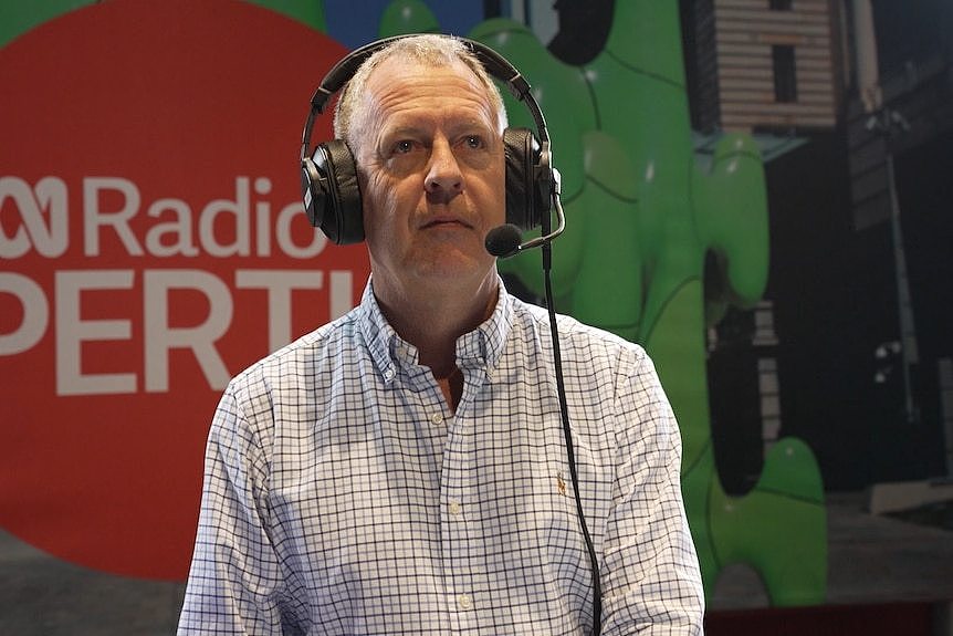 A man in a business shirt stands in front of a sign that says ABC Radio Perth. He is wearing headphones that have a microphone.