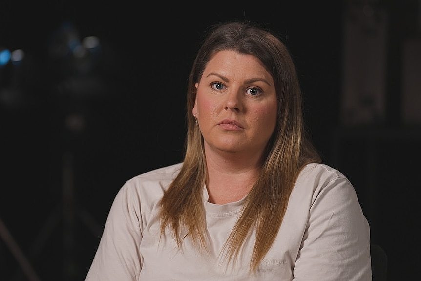 A woman sitting in a darkened room looks to the camera with a neutral expression.