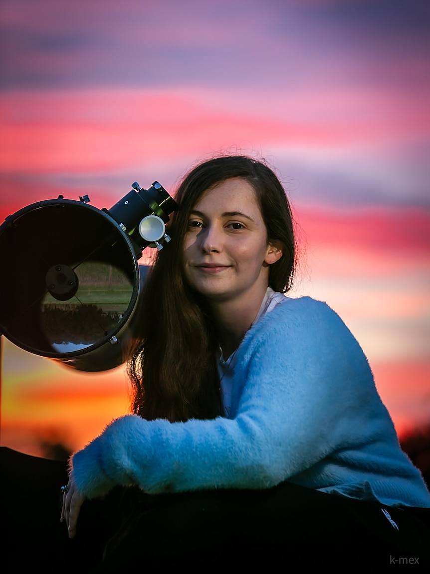 Krystal sits perched alongside a large telescope with a sunset sky behind her.