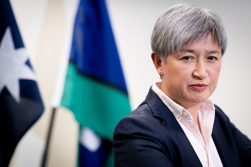 A close up shot of a woman with short grey hair in front of flags. 