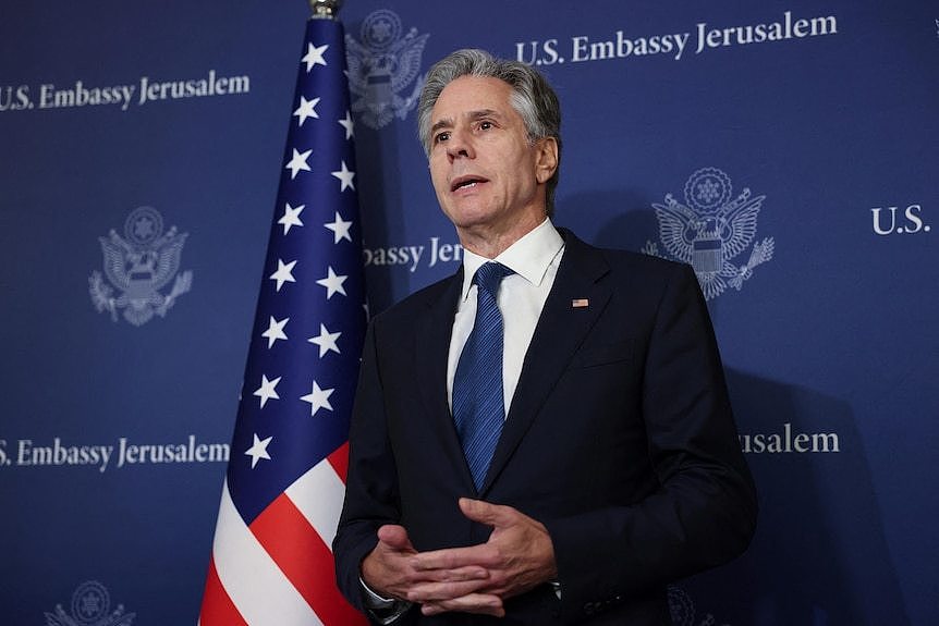 A man wearing a black suit and blue tie standing next to the United States flag