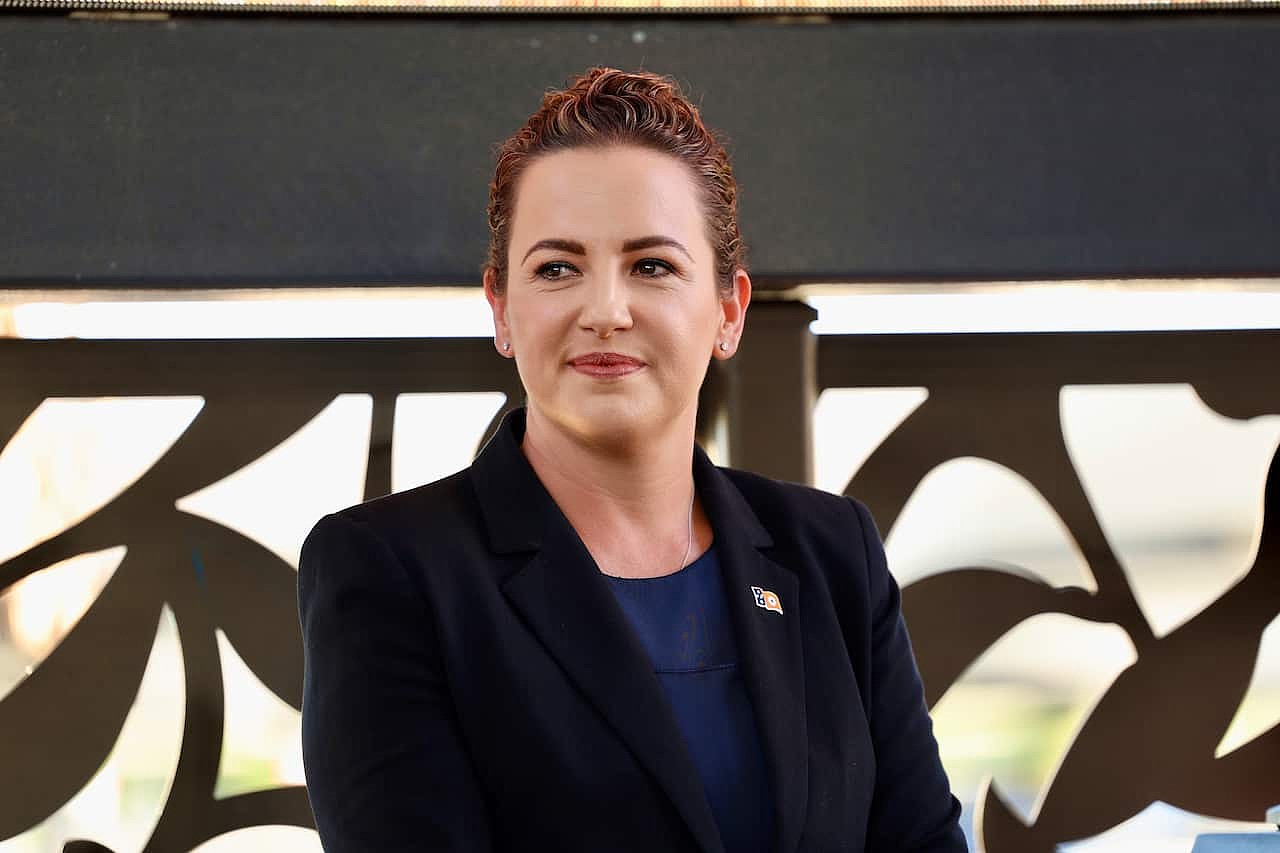 A woman with short red hair sitting down. She is wearing a blazer with a pin on it.