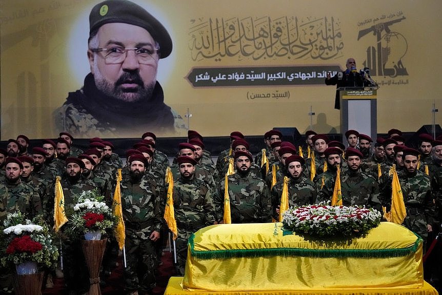 Fighters in camouflage stand in front of a coffin with a large picture of Fuad Shukr displayed on the wall behind