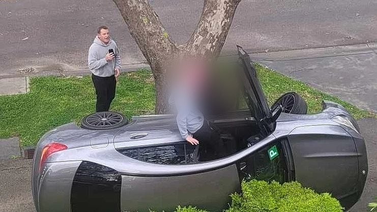 Michael Voss holds his phone up as two people scramble out of a Mercedes Benz crashed sideways in a residential street.