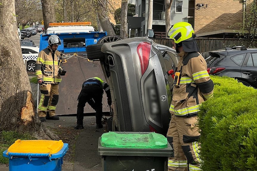 A crashed Mercedes Benz car on its side on a leafy street, as emergency crews look on.