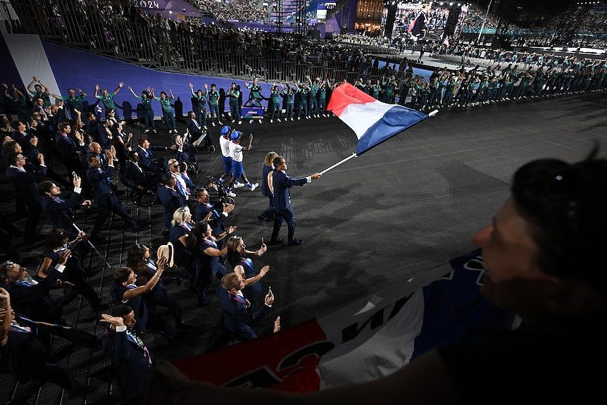 A French athlete waves the French flag as he and a teammate lead the team toward a big crowd and a stage at the Paralympics.