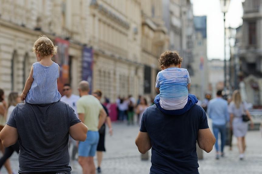 Two men walking on a busy street with children on their shoulders.