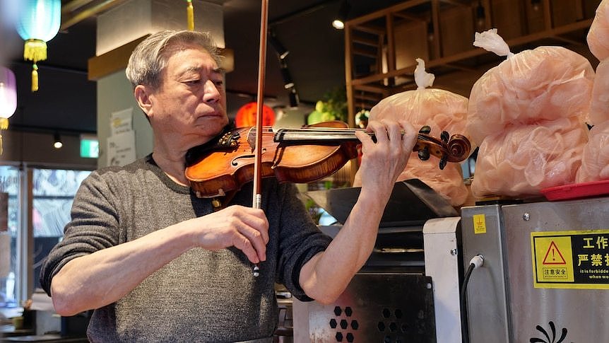 a man plays violin inside a restaurant next to three bags of pink prawn crackers