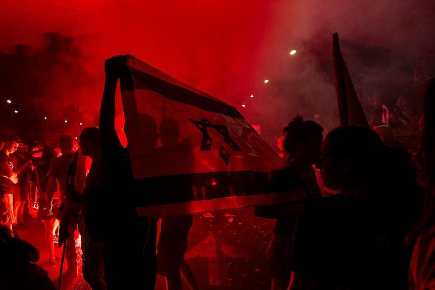 A group of young men and women waving an Israeli flag in the street are silhouetted against red light from a fire.