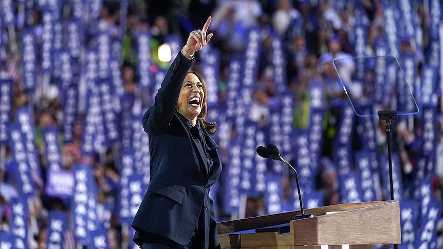 Kamala Harris smiles and points at something above her in the distance, in front of a crowd.