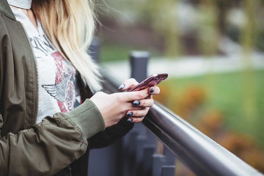 A girl with long hair is seen holding a phone while standing near a balcony overlooking a garden