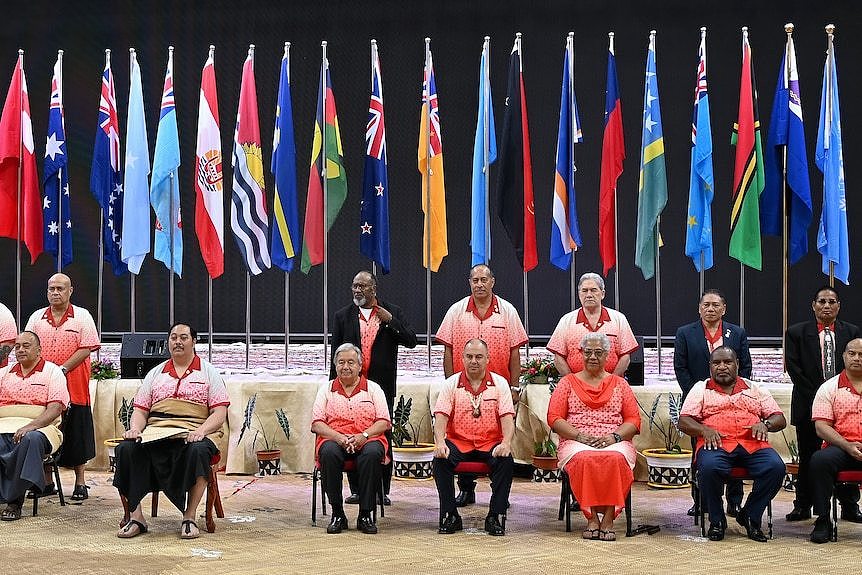 A group poses for a photo on chairs in front of a row of national flags.