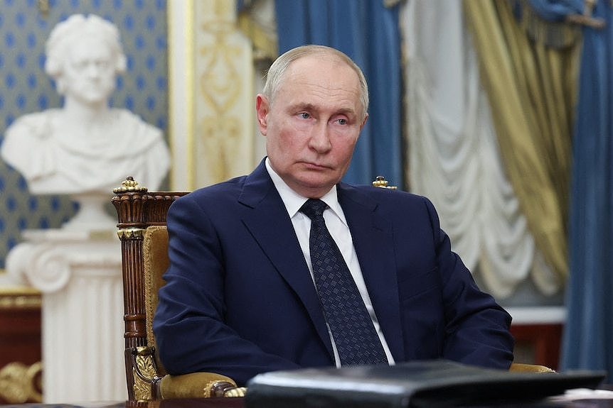 Vladimir Putin sits in a seat thinking while wearing a dark suit.