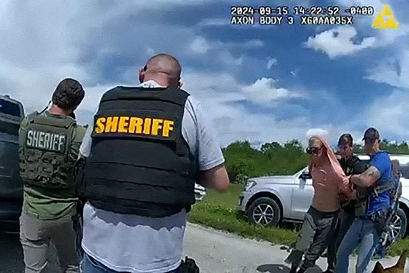 A man in a pink shirt is surrounded by police in flak jackets 