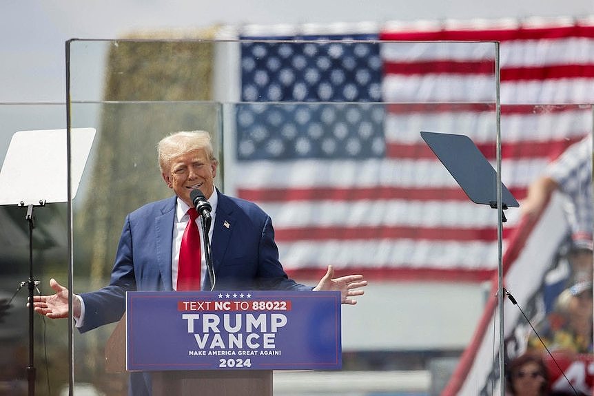 Donald Trump stands behind bullletproof glass onstage at an outdoor rally.