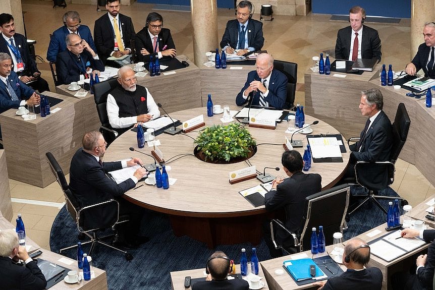 Quad members including Joe Biden and Narendra Modi sit at a round table, surrounded by men in suits.