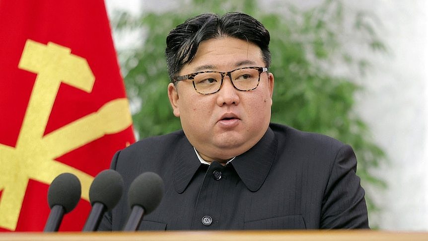 Kim Jong Un speaks in front of microphones with a red and yellow flag in the background 